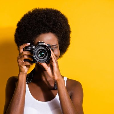 Black woman with camera up to eye on yellow background